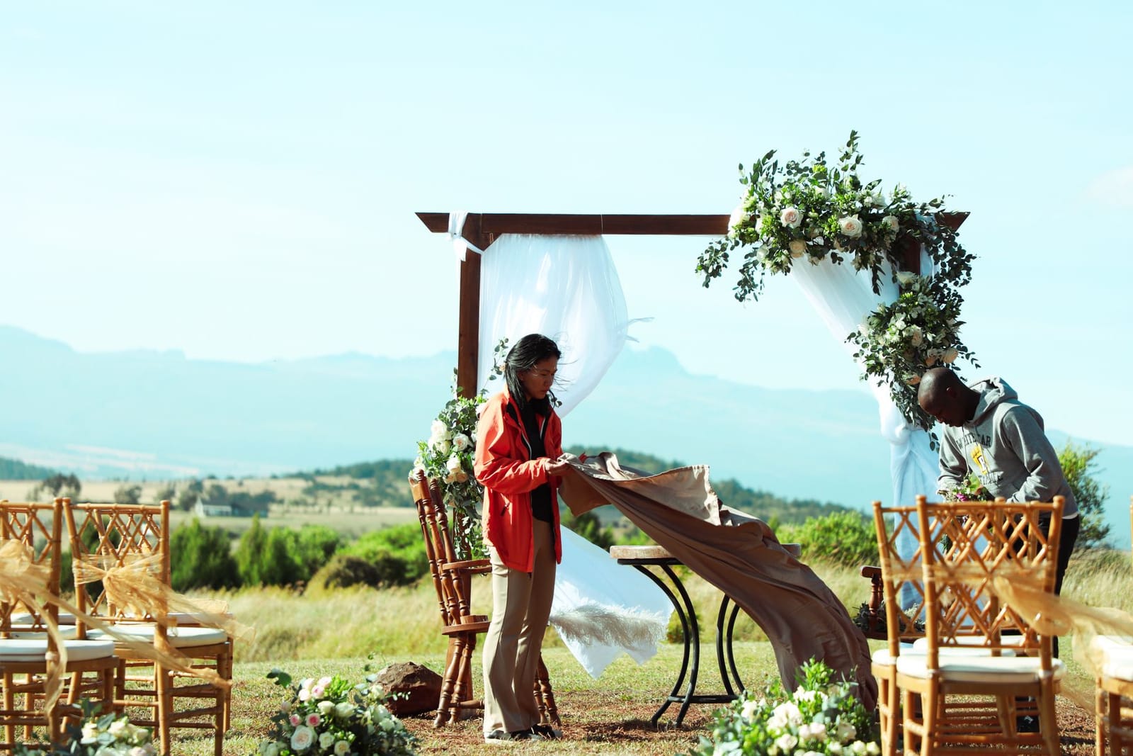 A woman in a red jacket lays a runner over a table beside a wooden ceremony arch while a man adjusts the flowers