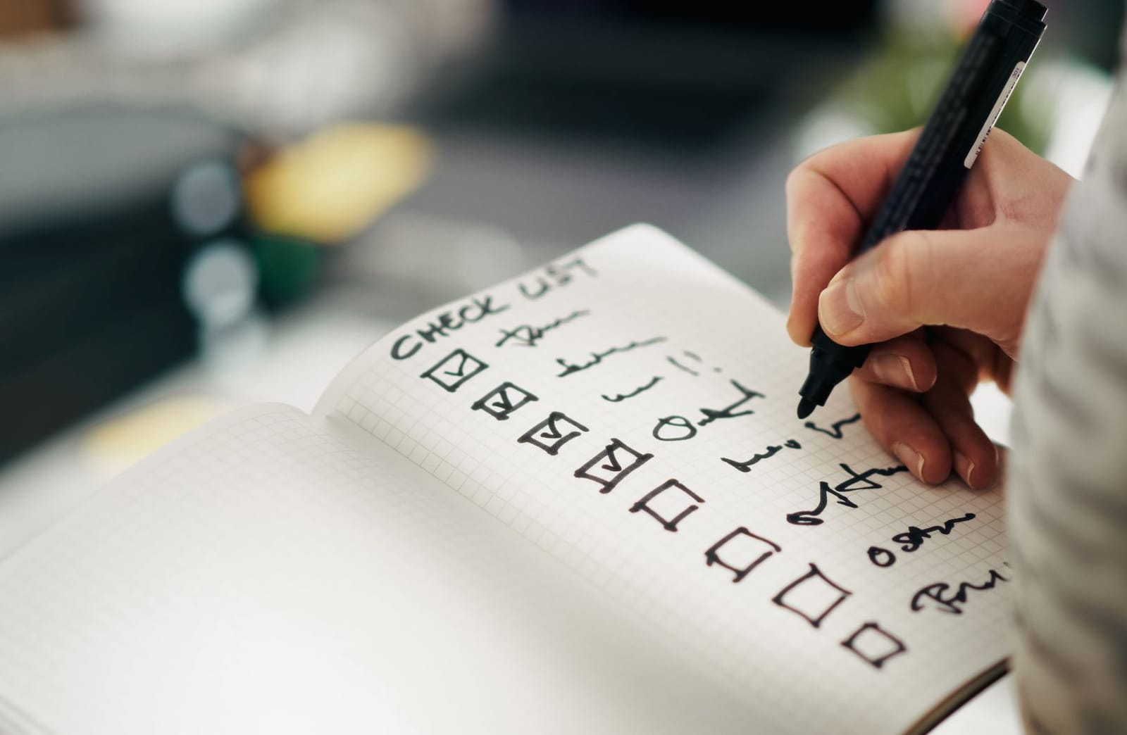 A hand ticks items on a handwritten checklist in a squared notebook