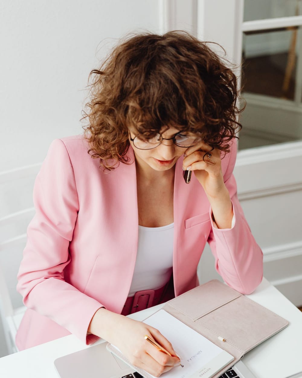 A woman in a pink blazer takes notes in a notebook while on the phone, a laptop open in front of her