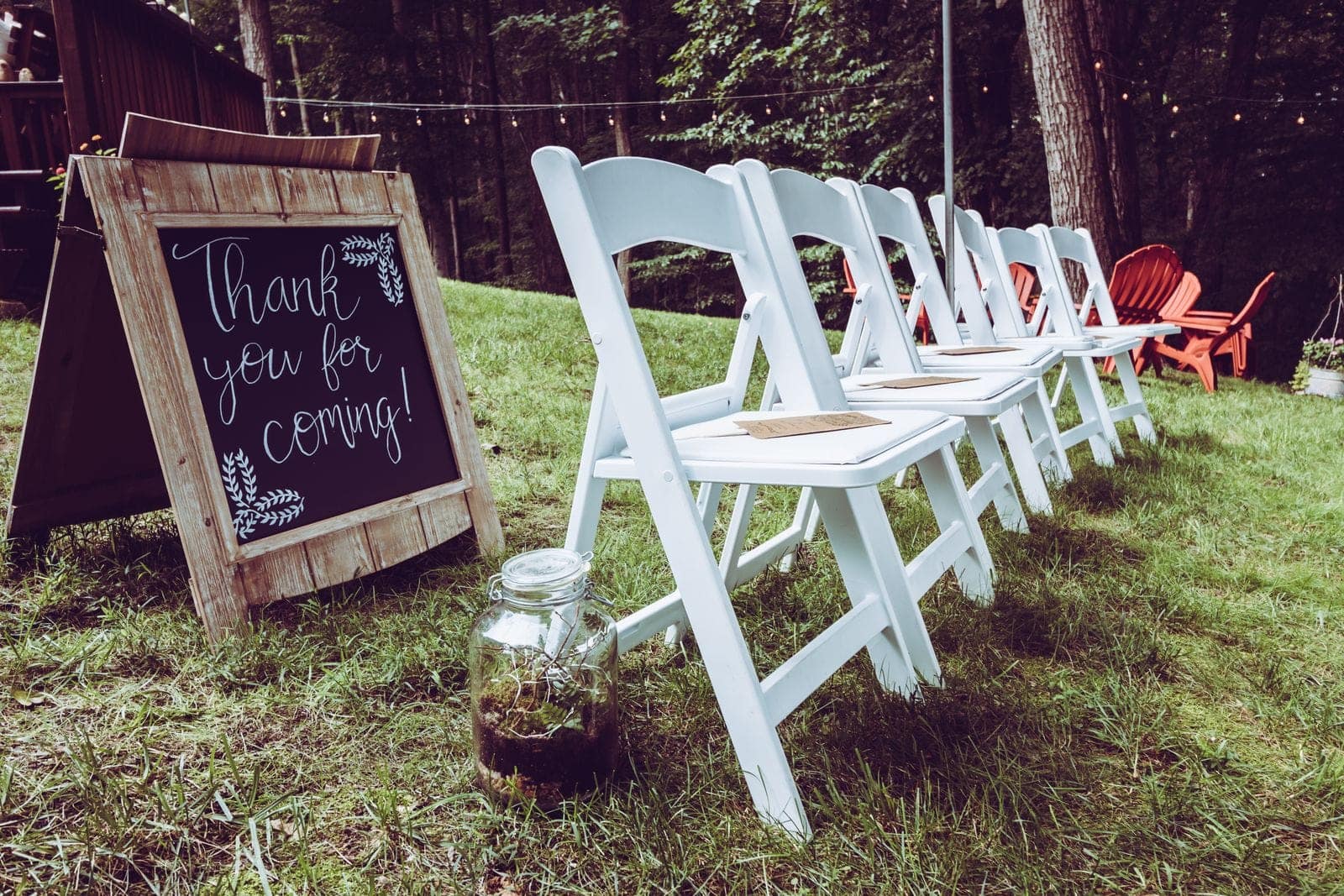 White folding chairs on a lawn beside a hand-lettered "Thank you for coming!" chalkboard, set for a backyard ceremony