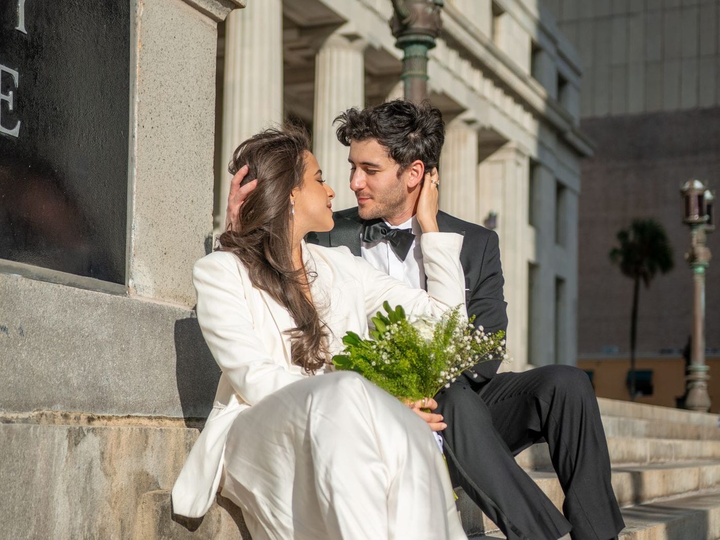 A couple in wedding clothes sitting on the stone steps of a civic building, holding a small bouquet