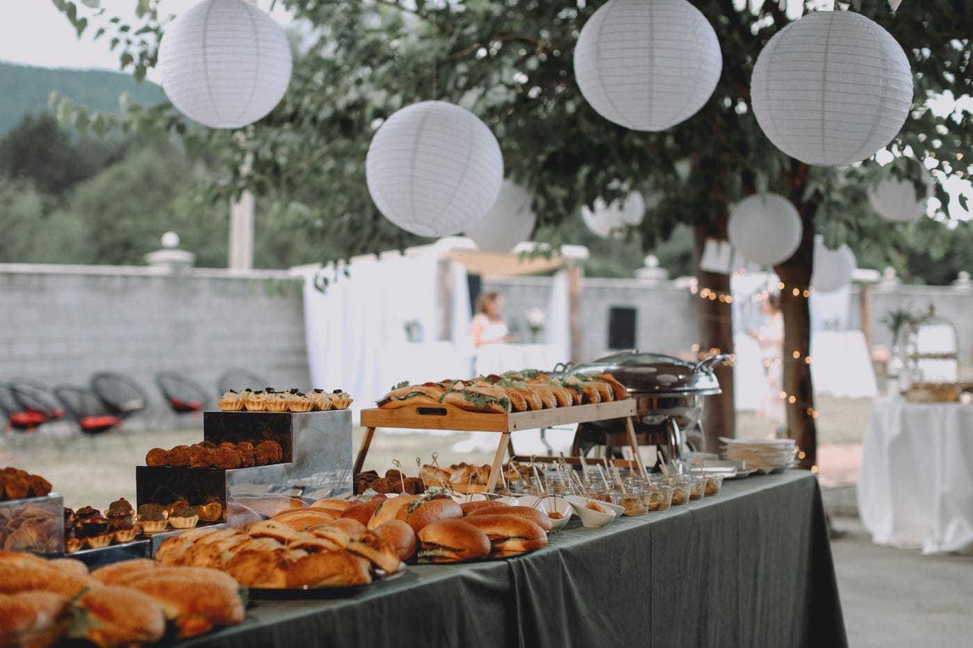 An outdoor buffet table of sandwiches and pastries under white paper lanterns