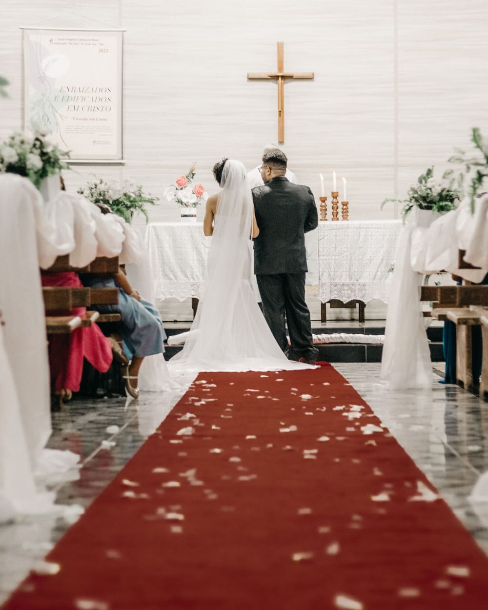 Back view of a couple at a church altar under a wooden cross, on a red runner scattered with petals