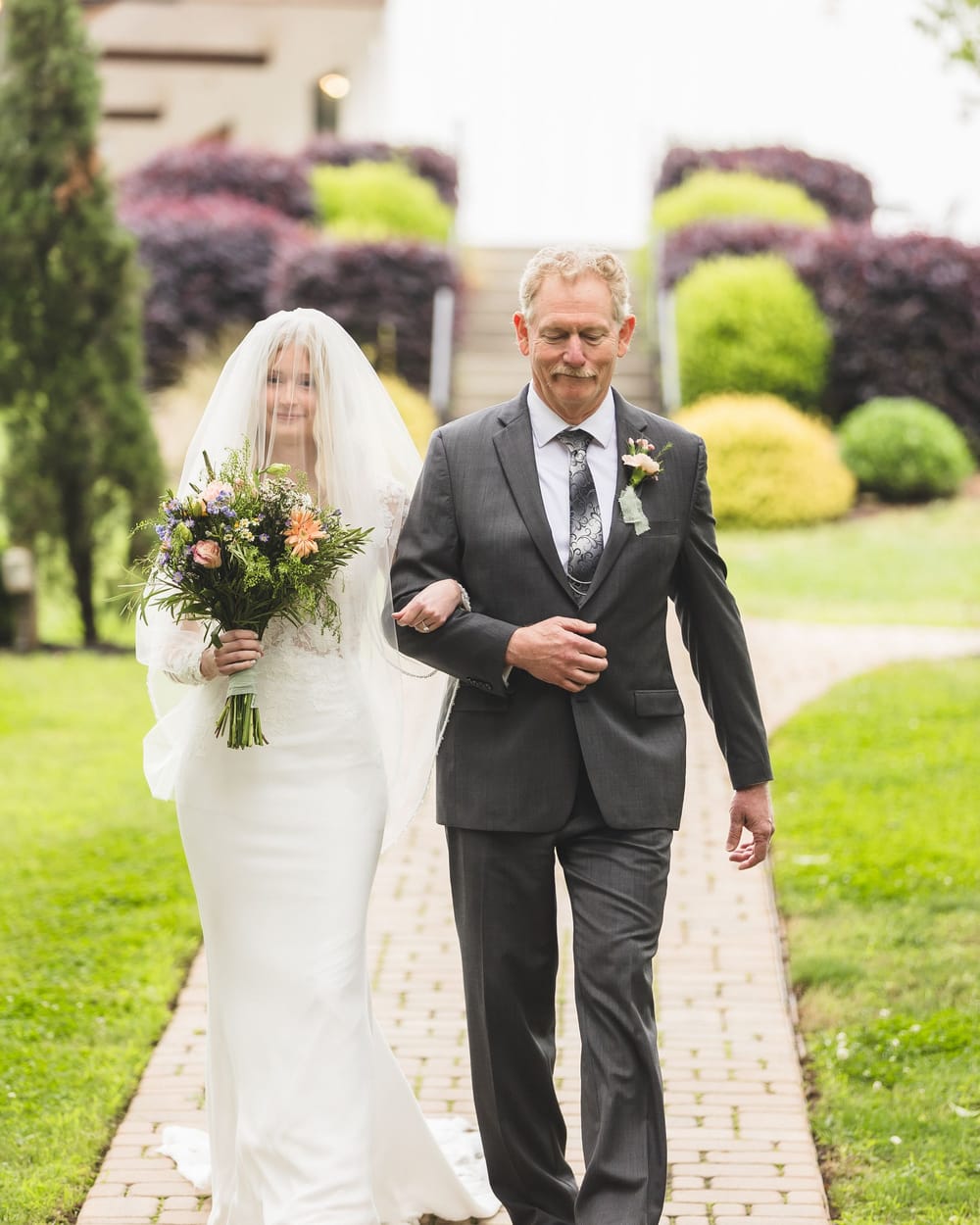 A veiled bride holding a wildflower bouquet walks a brick path on an older man's arm
