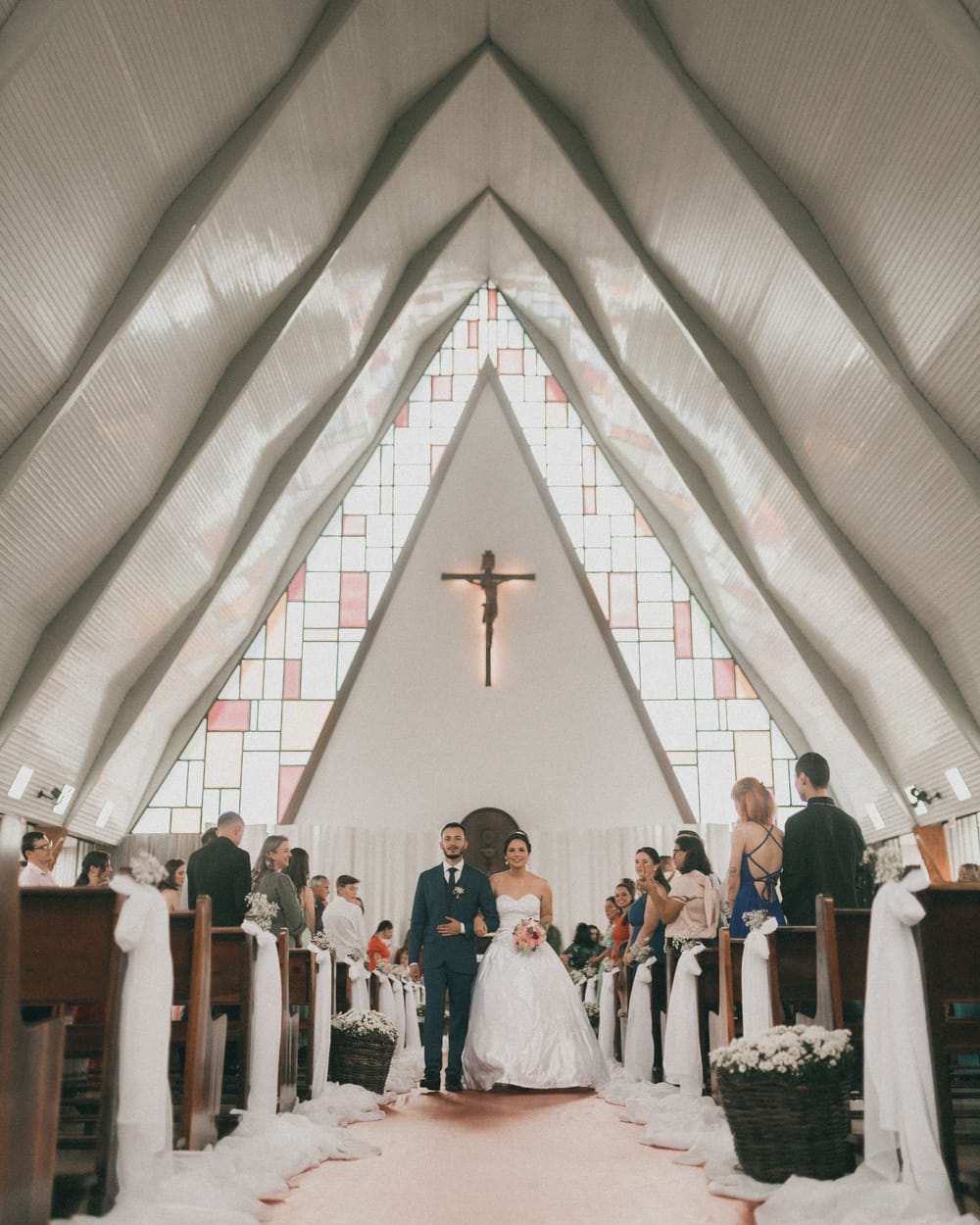 A couple walks back up the aisle of a church with a peaked roof and a stained-glass wall as guests stand