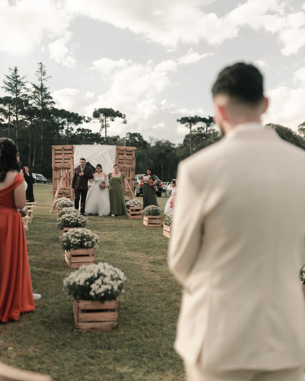 A bride walks a grass aisle lined with flower crates between a man and a woman, toward a man waiting at the front