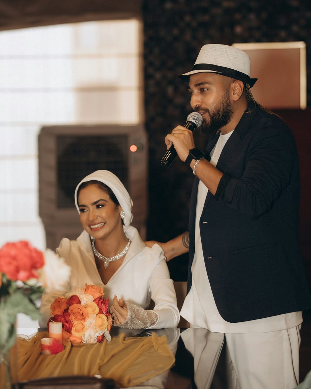 A groom in a white hat speaks into a microphone while his bride laughs beside him at the table