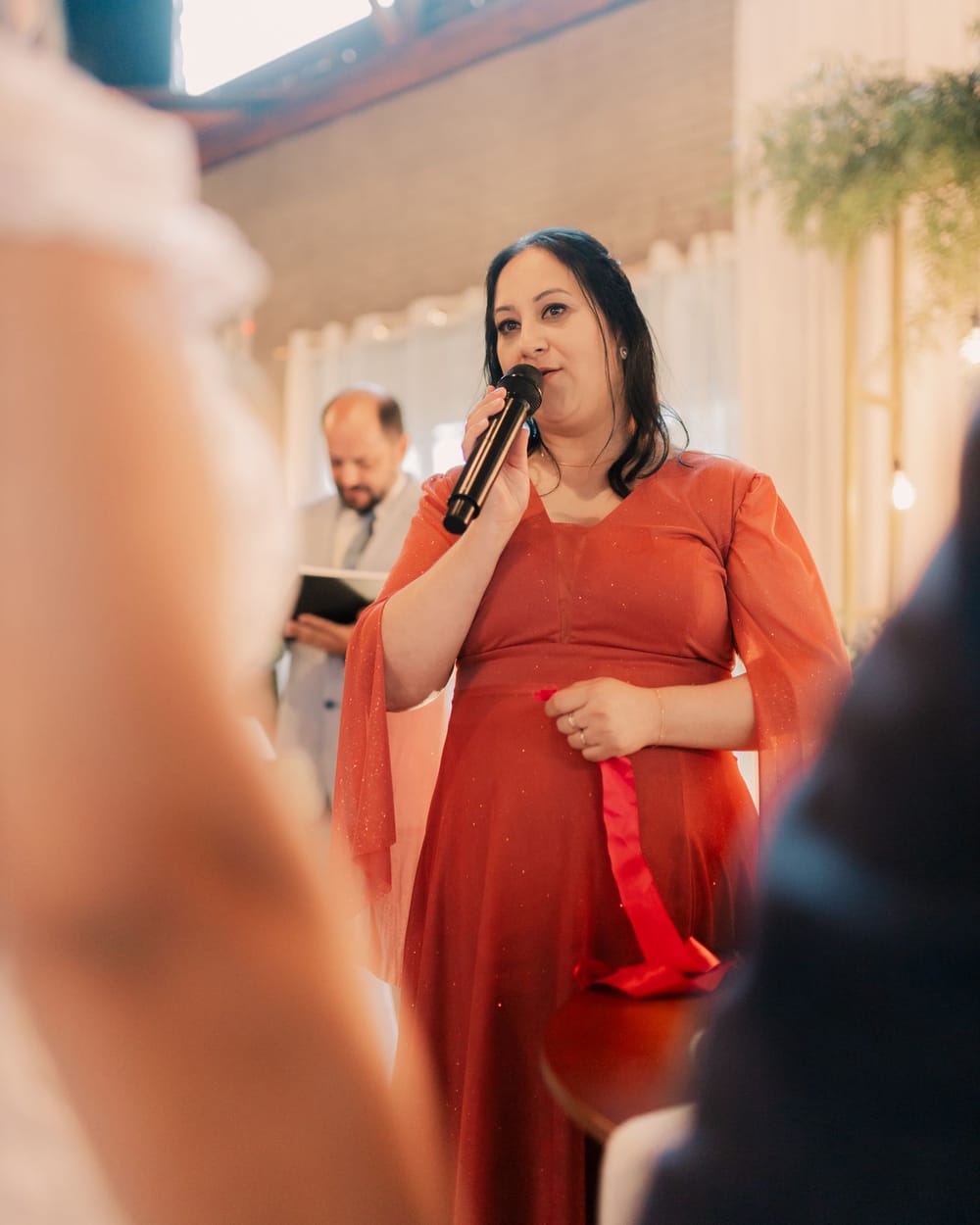 A guest in a rust-colored dress speaks into a microphone, the officiant standing behind her