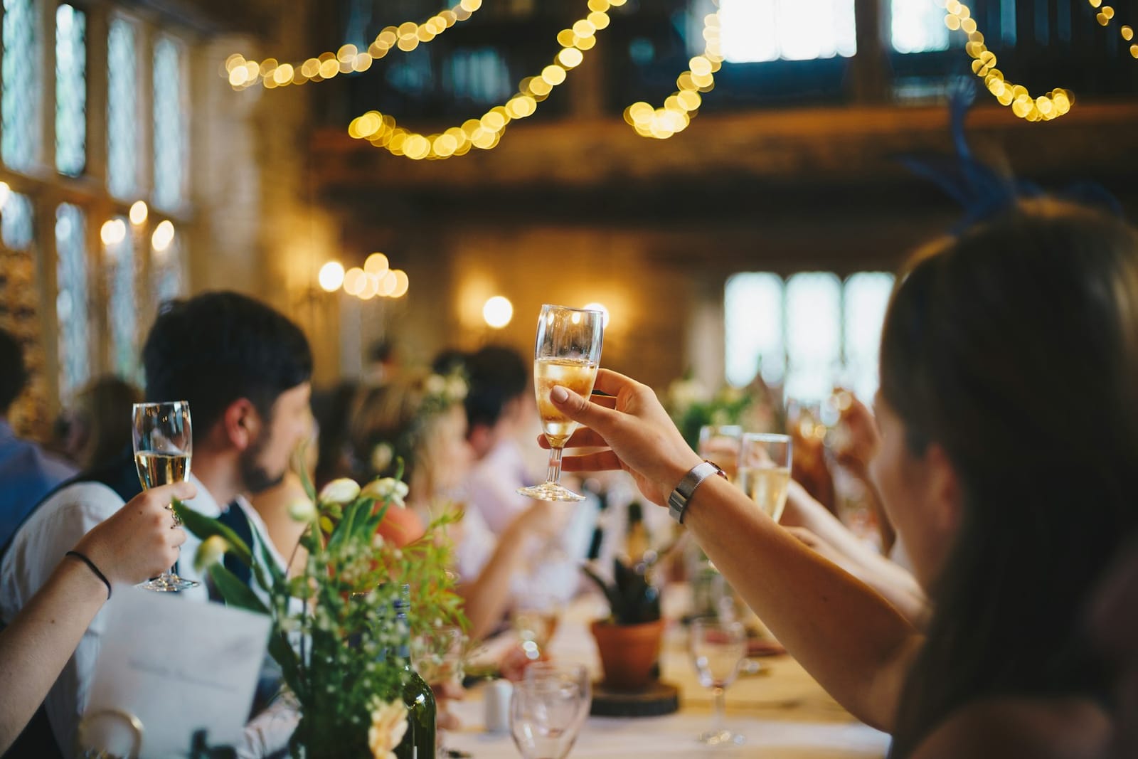 Guests raise glasses of champagne at a long reception table under string lights