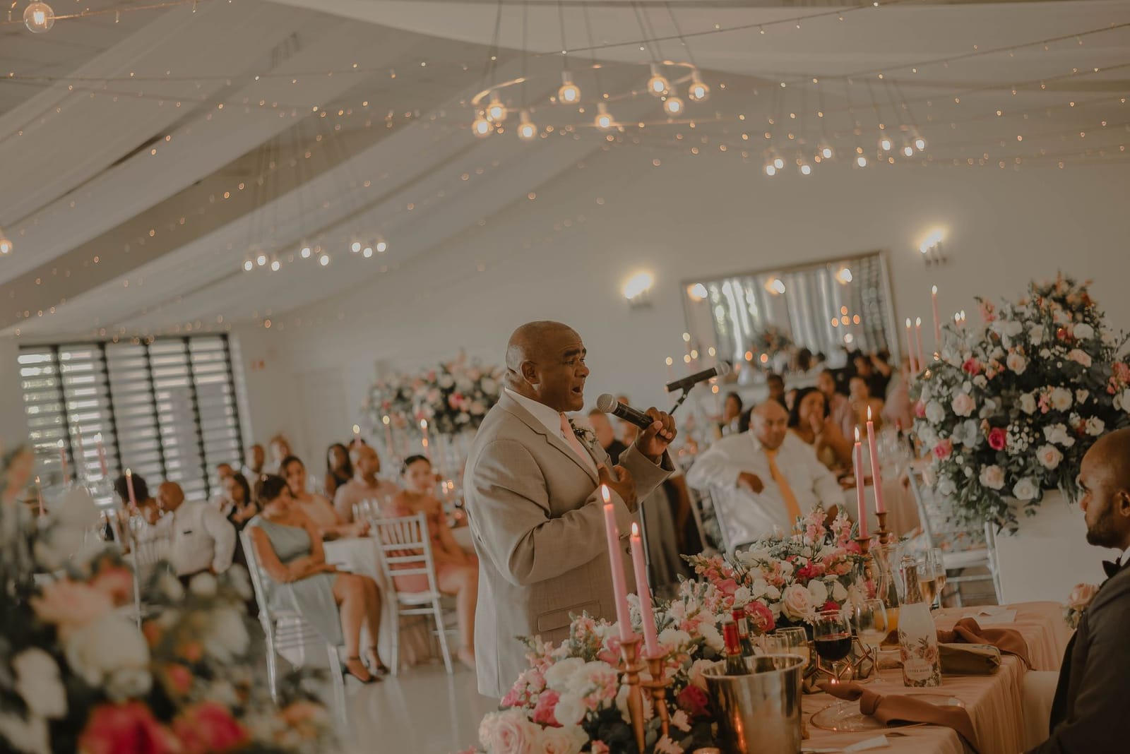 A man in a beige suit speaks into a microphone at the head table of a candlelit reception