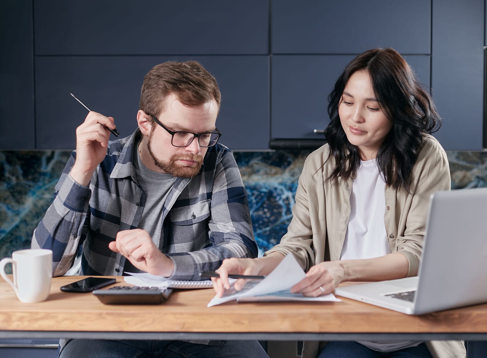 A man and a woman at a table with papers, a calculator, a phone and an open laptop