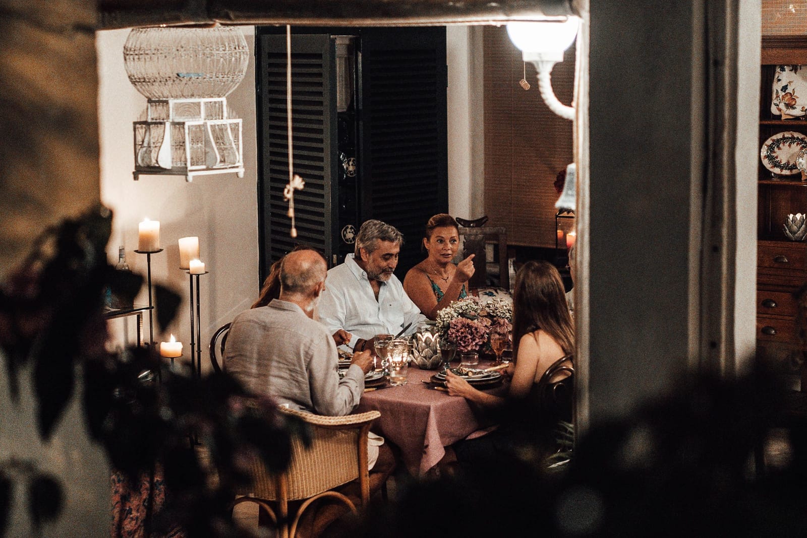An evening dinner seen through a doorway: five adults at a candlelit table with a pink cloth and flowers