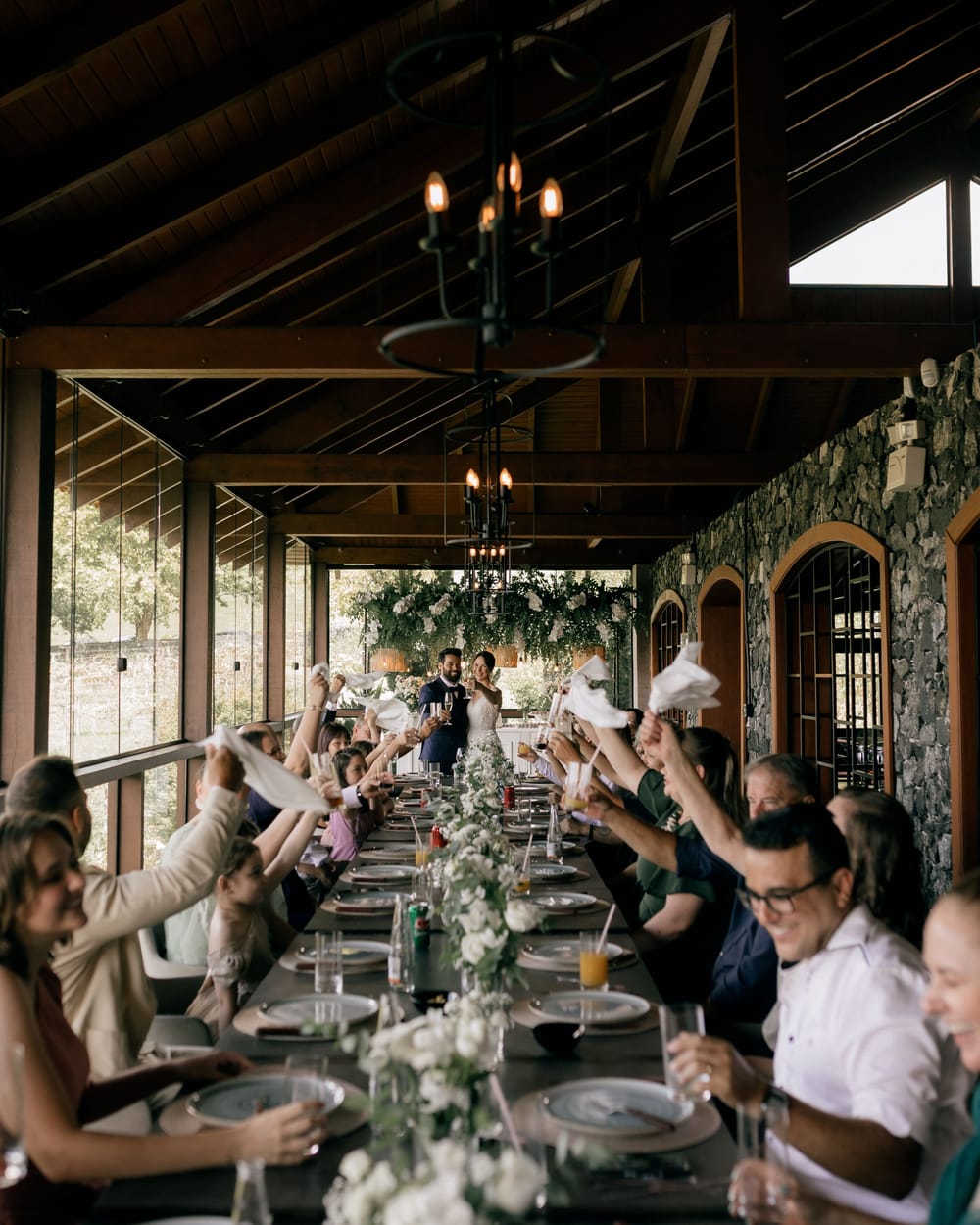 One long reception table under a wooden roof with chandeliers, guests on both sides waving white napkins