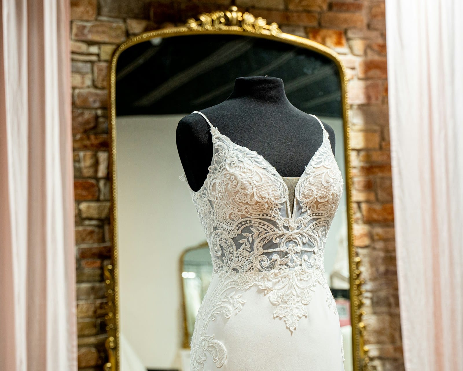 A lace wedding gown on a dress form in front of a gold-framed mirror in a bridal boutique