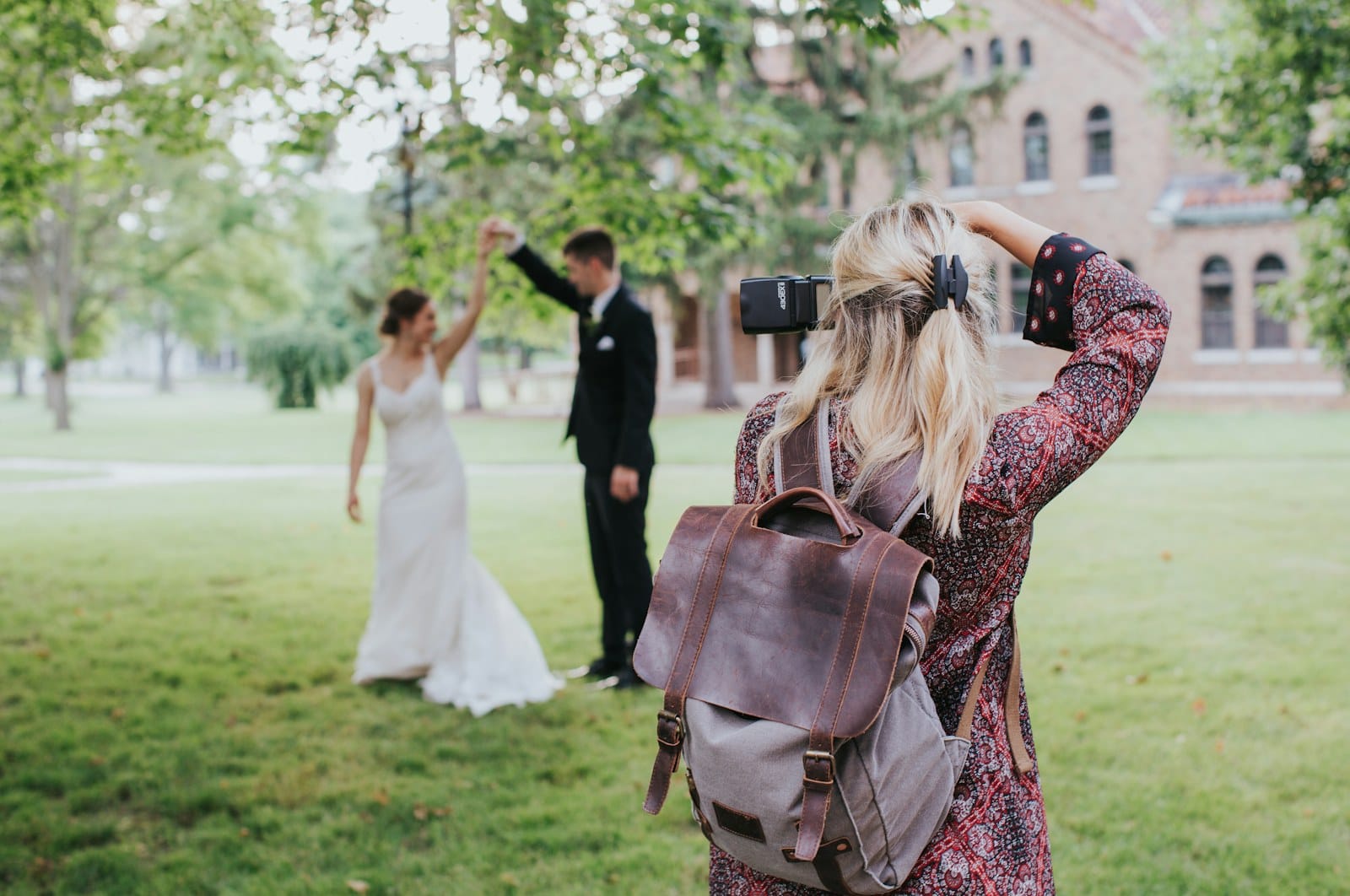 A wedding photographer with a leather backpack photographs a couple dancing on a lawn