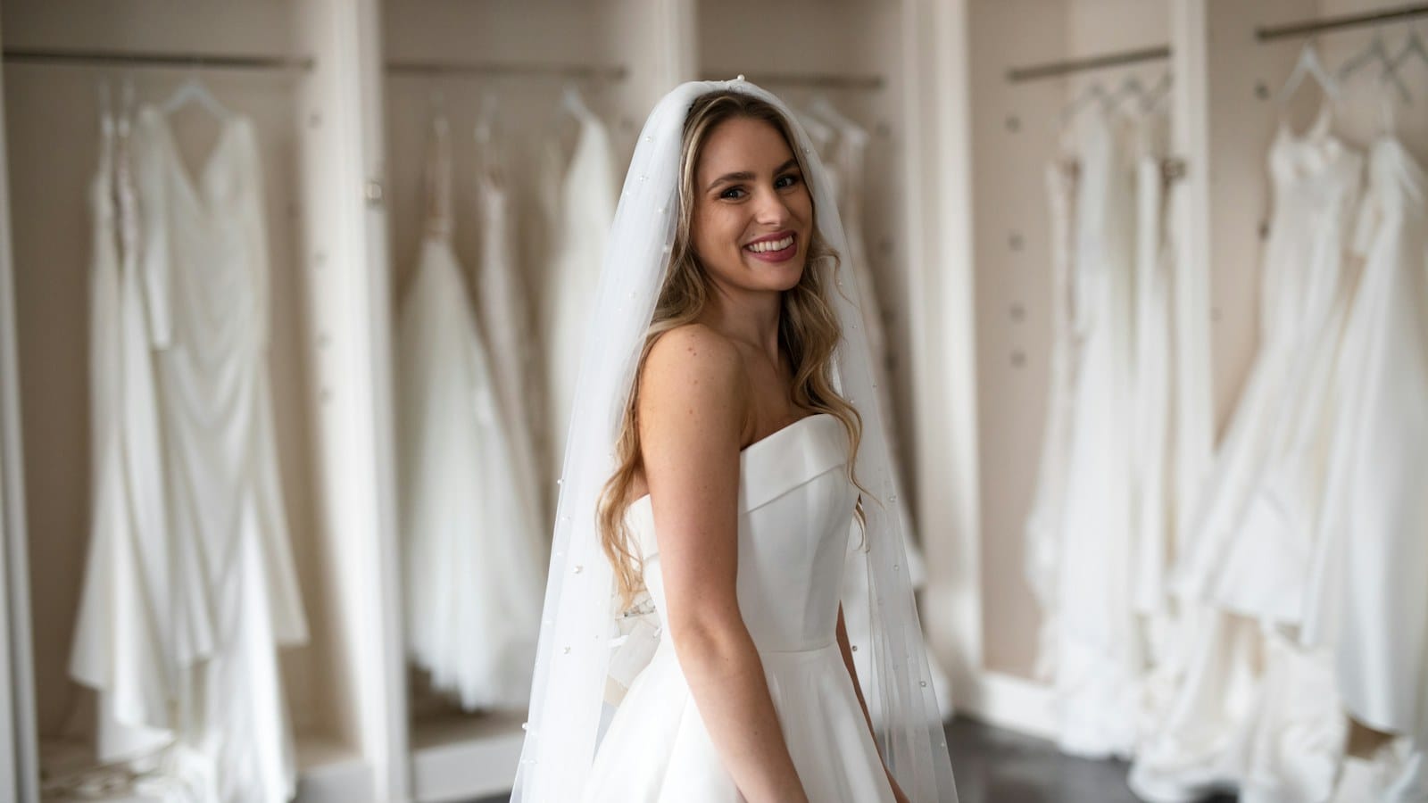 A bride in a strapless gown smiling in a bridal shop, racks of wedding dresses behind her
