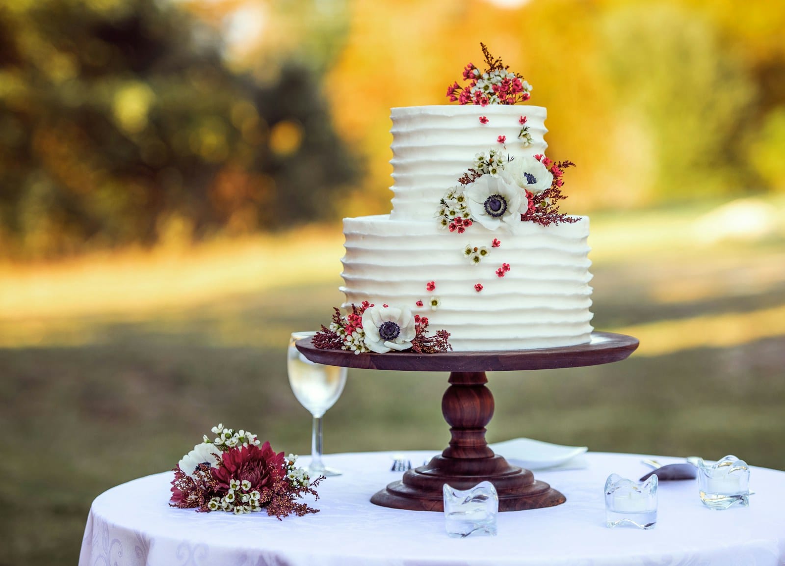 A two-tier white wedding cake decorated with red and white flowers on a wooden stand outdoors