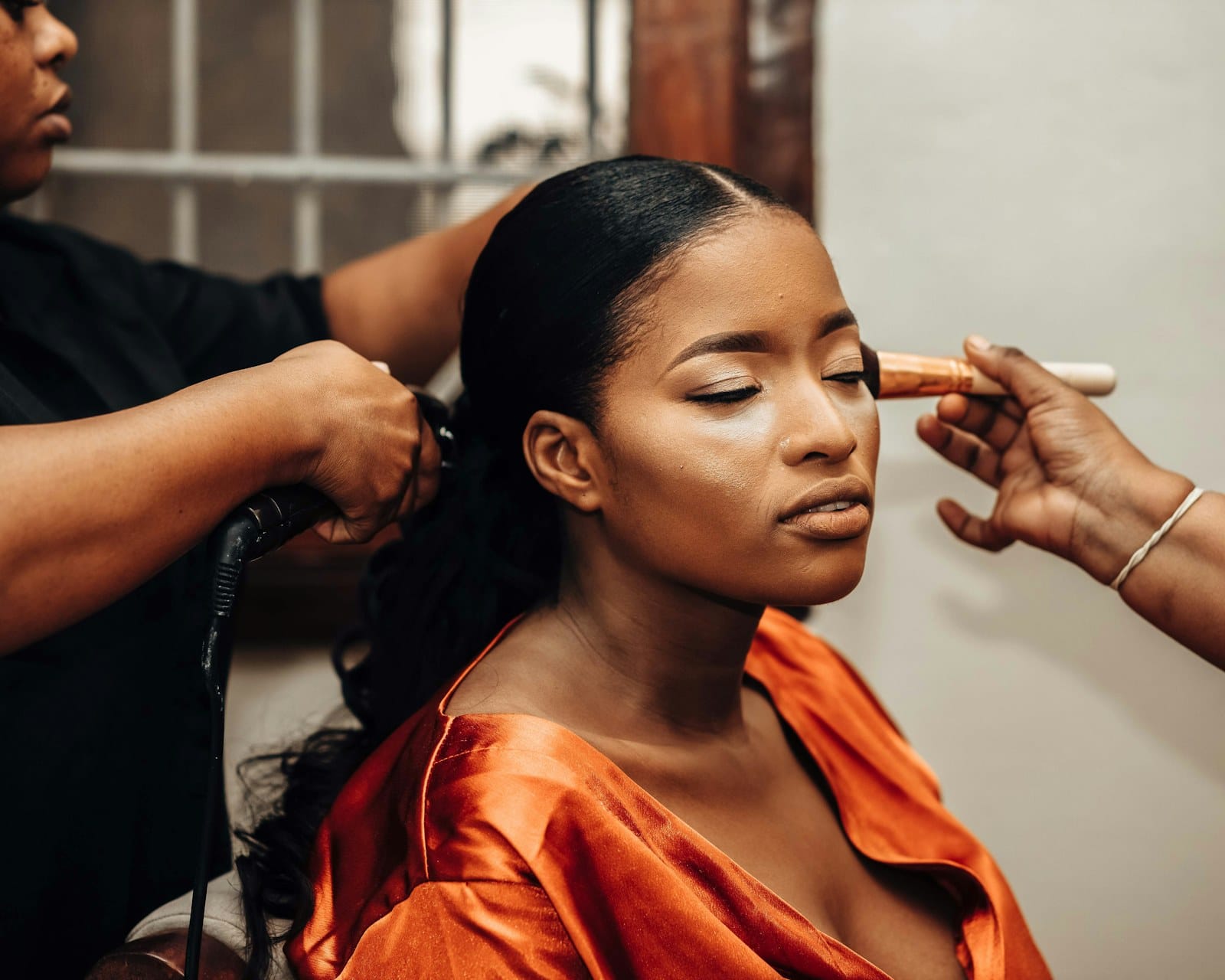 A bride in an orange robe having her hair styled and her makeup applied at the same time